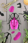 Pink paper violin beetle craft by Assembli featuring folded paper wings and detailed body on a plain background.