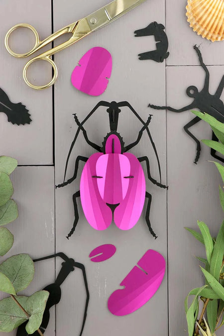 Pink paper violin beetle craft by Assembli featuring folded paper wings and detailed body on a plain background.
