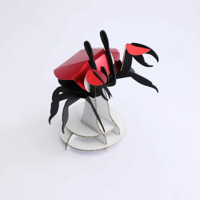 Colorful paper beach crab craft by Assembli, featuring bright red and orange paper legs and claws on a sandy background.