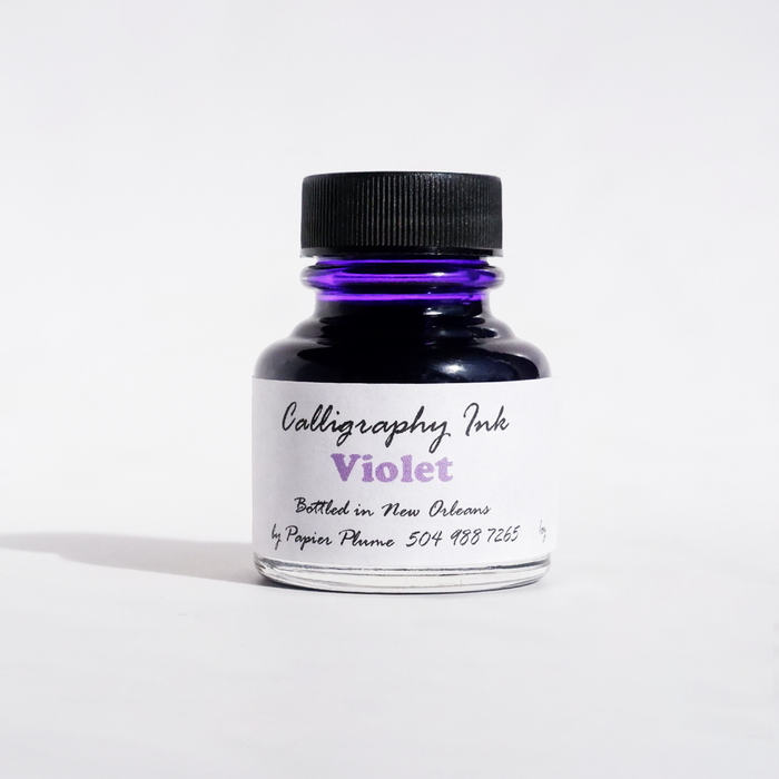 Violet bottled calligraphy ink by Papier Plume with a glass ink bottle and calligraphy pen on a white background.